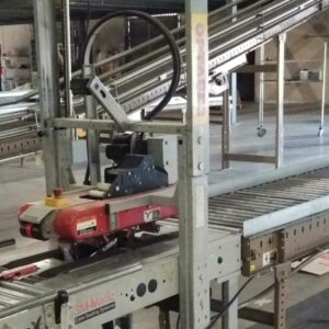 Close-up view of an industrial case sealing machine in a warehouse setting. The machine features a red sealing unit mounted on a metal frame with conveyor belts running through it. Surrounding the machine are various other conveyor systems and industrial equipment. The environment appears functional and organized, indicating a well-maintained and operational facility.