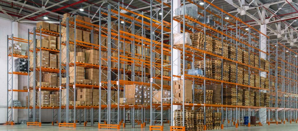 A spacious warehouse interior features tall, multi-level pallet rack shelving units filled with neatly stacked cardboard boxes and wooden pallets. The pallet rack shelves are constructed with grey vertical frames and bright orange horizontal supports, creating a vibrant and organized appearance. The pallet rack shelving units extend high towards the ceiling and run the entire length of the warehouse, showcasing the extensive storage capacity. The warehouse floor is clean and polished, with ample space for movement and operations. The high ceiling is supported by a network of exposed beams and industrial lighting, providing bright illumination throughout the space. The overall scene highlights a well-organized, efficient storage system, ready to support large-scale inventory management.