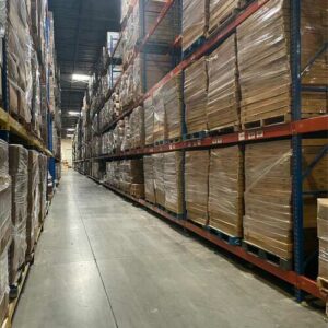 Interior of a warehouse featuring long aisles lined with tall metal pallet rack shelving units. The pallet rack shelves are stacked high with pallets of boxes wrapped in plastic, showcasing an extensive and well-organized inventory. The warehouse has a high ceiling with bright industrial lighting, providing clear visibility down the aisle. The concrete floor is clean, and the environment appears spacious and efficiently managed.