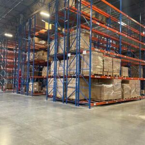 Interior of a spacious warehouse with tall metal pallet rack shelving units in blue and orange, filled with boxes and packages stacked on pallets. The pallet rack shelves are neatly organized, showcasing an efficient storage system. The warehouse has a high ceiling with bright industrial lighting, providing clear visibility throughout the space. The concrete floor is clean and the overall environment appears well-maintained and orderly.