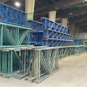 A spacious warehouse interior featuring neatly stacked pallet rack uprights in blue and green. The uprights are organized into multiple tiers, creating an orderly storage area along both sides of a wide aisle. The high ceiling with exposed beams and bright overhead lighting provides ample illumination. The floor is clean and clear of debris, indicating a well-maintained and efficient storage environment. The overall scene highlights the organized storage solution for pallet racking components within the warehouse.