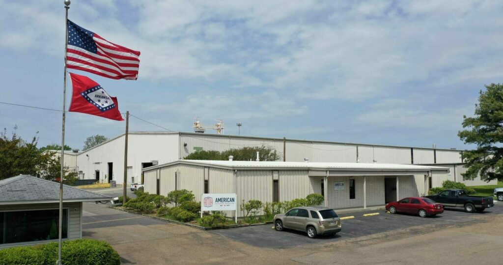 Exterior view of American Material Handling headquarters with an American flag and an Arkansas state flag flying on flagpoles in the foreground. The building is a large, beige-colored warehouse with a smaller attached office section. There are a few cars parked in the lot in front of the office. The scene is set on a partly cloudy day, with trees and shrubs adding greenery around the premises.