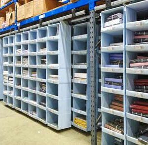 A well-organized Storeganizer warehouse section features multiple shelving units filled with light blue storage compartments. Each compartment holds neatly stacked items, such as folded fabrics or garments, sorted by color and type. The shelving units have grey metal frames and blue horizontal supports, providing a sturdy structure for the compartments. Above the compartments, larger boxes are stored on higher shelves, adding to the efficient use of vertical space. The concrete floor is clean and clear, allowing easy access to the items. The overall scene reflects a highly organized storage system designed for easy identification and retrieval of inventory.