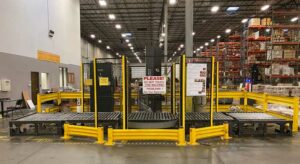 Interior of a large warehouse featuring a used auto pallet wrap machine with yellow safety guard rails. The auto pallet wrap machine is surrounded by a wire modular partition safety barrier with a sign that reads, "PLEASE! DO NOT TOUCH THIS MACHINE! PROBLEMS? See Your Supervisor." The background shows tall pallet rack shelving units stocked with various items. Bright industrial lighting illuminates the clean and well-maintained environment.