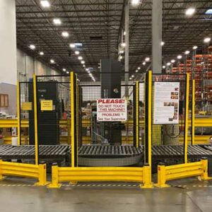 Interior of a large warehouse featuring a used auto pallet wrap machine with yellow safety guard rails. The auto pallet wrap machine is surrounded by a wire modular partition safety barrier with a sign that reads, "PLEASE! DO NOT TOUCH THIS MACHINE! PROBLEMS? See Your Supervisor." The background shows tall pallet rack shelving units stocked with various items. Bright industrial lighting illuminates the clean and well-maintained environment.