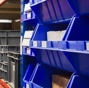 A close-up view of warehouse shelving units filled with blue and grey plastic storage bins. The blue bins are prominently displayed in the foreground, neatly arranged on shelves with white labels for identification. The grey bins are visible on adjacent shelves, also well-organized. The shelving units have blue vertical frames and red horizontal supports, creating a sturdy structure. The background shows more storage bins and items, indicating a well-maintained and efficient storage system designed for easy access and organization of small parts and inventory. The overall scene highlights the meticulous organization and effective use of space in the warehouse.