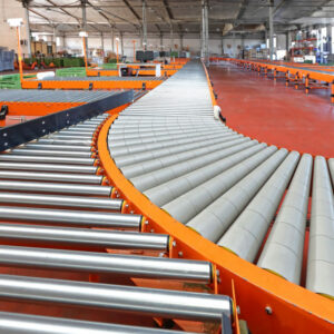 An industrial warehouse features a large powered conveyor system with shiny metal rollers and bright orange frames. The powered conveyor follows a smooth, curved path, allowing for efficient movement of goods around corners. The floor is painted a vibrant red, adding to the bright and organized appearance of the facility. In the background, various storage containers and equipment are visible, indicating a busy and well-organized working environment. The high ceiling and spacious layout suggest a large-scale operation, with the conveyor system playing a crucial role in the material handling process. The overall scene highlights the efficiency and sophistication of automated logistics within the warehouse.