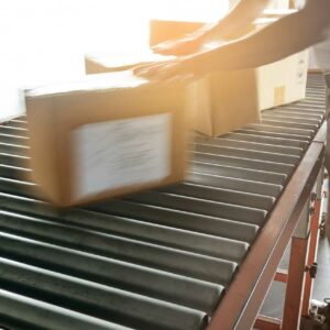 A close-up view of a person handling cardboard boxes on a conveyor system in a warehouse. The conveyor consists of metal rollers and an orange frame, designed for efficient movement of packages. The person, dressed in khaki pants and a white shirt, is pushing a box along the rollers. The scene is brightly lit, with sunlight streaming in from the left, creating a warm and dynamic atmosphere. The overall image highlights the hands-on process of package handling and the role of conveyor systems in facilitating smooth and efficient material flow in a warehouse setting.