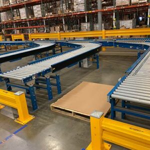 A warehouse interior features a used powered conveyor system with metal rollers and sturdy blue frames. The conveyor has a curved section, allowing it to navigate turns efficiently. Bright yellow safety barriers are installed around the conveyor to protect equipment and personnel. In the foreground, a wooden pallet with a large sheet of cardboard is placed on the floor. The background shows rows of tall shelving units filled with various boxes and inventory, indicating a well-organized storage area. The clean, polished concrete floor and the systematic arrangement of the conveyor system highlight the facility's focus on efficiency and safety in material handling operations.