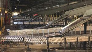A large industrial facility features an extensive network of gravity conveyors. The foreground shows a series of flexible, accordion-like conveyor sections on wheels, designed for easy maneuverability and adjustment. Overhead, additional conveyor belts are visible, creating a multi-level system for efficient material handling. The facility has a high ceiling with visible structural supports and industrial lighting, emphasizing its scale and capacity for high-volume sorting and distribution operations. The overall scene highlights a well-coordinated, automated process essential for large-scale logistics and warehousing.