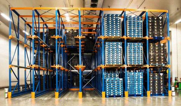 The image shows a high-density drive-in pallet rack system in a warehouse. The drive-in pallet racks are constructed with blue vertical uprights and orange horizontal beams, creating a multi-level storage solution. Each level is filled with stacked blue and black pallets containing various items, indicating efficient use of vertical space. The pallets are neatly organized, allowing for easy access and inventory management. The warehouse is well-lit with bright ceiling lights, and the concrete floor is clean and polished. The overall setup suggests a robust and organized system for storing large quantities of goods, likely for distribution or long-term storage.