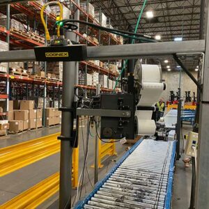 A warehouse setting features a used Cognex automated labeling machine mounted on a metal frame above a conveyor system with metal rollers and a blue frame. The machine is equipped with rolls of labels and various electronic components for automated operation. In the background, tall shelving units filled with boxes and inventory are visible, along with bright yellow safety barriers. The warehouse has a high ceiling with industrial lighting, providing ample illumination. A worker wearing a high-visibility vest can be seen in the distance, indicating an active and organized workspace focused on efficient material handling and processing