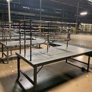 Interior of a large warehouse with several used long metal packing tables arranged in rows. The packing tables have multiple tiers of shelving above the main tabletop, providing ample storage and workspace. The area is well-lit with industrial lighting, and the concrete floor is clean. In the background, more packing tables and equipment can be seen, indicating a spacious and organized work environment.