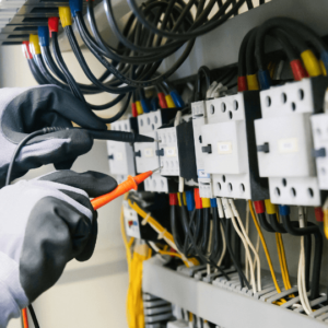A close-up of an electrician's hands, wearing gray gloves, working on electrical wiring in a control panel. The electrician is using testing probes on the wiring and connections. Various colored wires and electrical components are visible in the panel