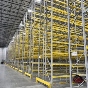 A spacious warehouse interior is shown with tall metal pallet rack shelving units arranged in long rows. The pallet rack shelving units feature bright yellow horizontal supports and grey vertical frames, creating a vibrant and organized appearance. The pallet racks extend high towards the ceiling and deep into the distance, showcasing the warehouse's extensive storage capacity. The concrete floor is clean and polished, reflecting the overhead industrial lighting. Safety barriers in matching yellow are positioned at the base of the pallet rack shelves for protection. A red tool bag is visible on the floor in the foreground, adding a touch of color and indicating an active work environment. The overall scene highlights a well-organized and efficiently planned storage area, ready for inventory.