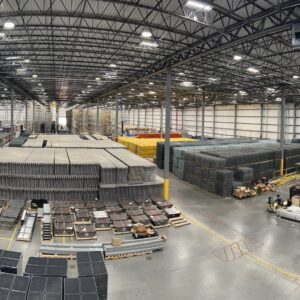 A panoramic view of a large, well-organized warehouse. The space is filled with neatly stacked pallets, various types of industrial materials, and assembled racking systems. Overhead, a network of metal trusses supports bright lighting, illuminating the entire area. There are designated areas for different materials, including a section with yellow and gray pallets stacked high. In the foreground, workstations and pallets with organized components are visible, along with a clear pathway for movement. The warehouse features high ceilings and ample space, indicating a well-maintained and efficiently run facility.