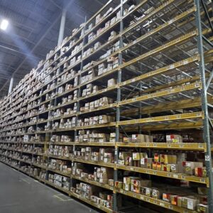 A large warehouse aisle features extensive shelving units filled with numerous cardboard boxes. The shelves, made from sturdy metal frames with yellow supports, rise high towards the ceiling, showcasing the warehouse's significant storage capacity. Each shelf is densely packed with boxes of various sizes, each marked with colorful labels and tags for inventory identification. The shelving extends far into the distance, emphasizing the scale of the storage area. The warehouse floor is clean and orderly, with clear pathways for movement.