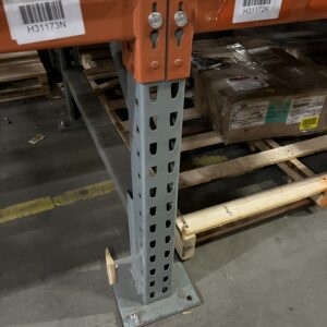 Close-up view of the base of a metal shelving unit in a warehouse. The shelving unit has gray uprights and orange horizontal beams with wire mesh decking. The upright is anchored to the concrete floor with a metal base plate and bolts. A labeled barcode is visible on the horizontal beam for organizational purposes. A pallet with a cardboard box is seen on the lower shelf, and the surrounding floor area is clean and well-maintained.