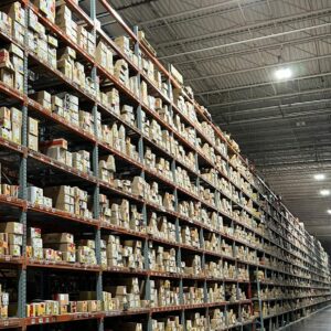 A vast warehouse aisle is shown lined with tall shelving units packed with numerous cardboard boxes. The shelves, constructed from sturdy metal frames with orange supports, extend high up towards the ceiling, filled with boxes of various sizes. Each box is labeled with multiple colorful tags and stickers, indicating inventory details. The shelving stretches far into the distance, demonstrating the extensive storage capacity of the warehouse. The warehouse floor is clean and organized, with clear walkways for easy access.