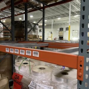 Close-up view of a partially stocked metal pallet rack shelving unit in a warehouse. The pallet rack shelves are orange with gray vertical uprights, and each shelf has labeled slots for organization. On the lower shelf, there are several large rolls of plastic wrap and other packaging materials. The background reveals more shelves and equipment in the spacious, well-lit warehouse. The overall setting is clean and organized, indicating an efficiently managed storage area.