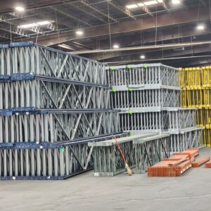 A well-organized warehouse with stacks of metal pallet rack shelving frames in gray, blue, and yellow colors. The pallet rack frames are arranged in large, orderly piles, with some orange metal beams stacked in the foreground. The warehouse has a high ceiling with industrial lighting, creating a bright and spacious environment. A broom is seen leaning against one of the stacks, indicating maintenance and cleanliness.