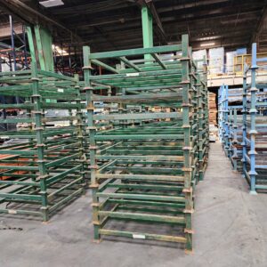 The image shows a collection of stack racks in a warehouse setting. These racks are made of metal and painted green, with some blue racks visible in the background. The stack racks are designed to be sturdy and durable, featuring multiple horizontal bars and vertical supports. They are stackable, allowing for efficient use of vertical space in the warehouse. The racks can be used for storing a variety of materials, such as pallets, containers, and other industrial goods. The warehouse environment includes other storage equipment and materials, with a concrete floor and high ceilings typical of industrial storage facilities. The overall setup highlights the organization and storage capacity of the warehouse