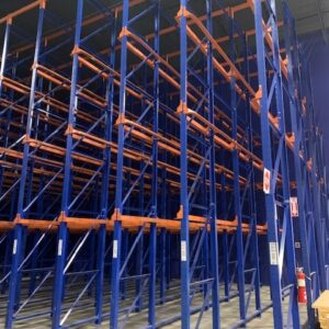 A warehouse interior features a tall, multi-tiered drive-in rack shelving system with blue metal frames and orange horizontal supports. The shelving units are extensive, forming a grid of tall, narrow aisles that reach up to the high ceiling. The floor is made of polished concrete, and the entire setup appears robust and designed for heavy-duty storage. Safety signs and a fire extinguisher are visible on one of the columns, indicating attention to safety regulations. The overall scene suggests a well-organized and spacious storage area, ready to hold a large volume of inventory.
