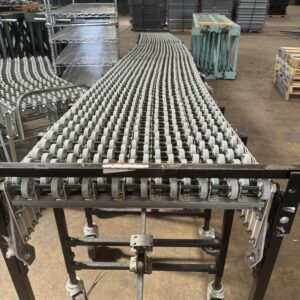 The image shows a Nestaflex conveyor system in a warehouse setting. The conveyor consists of multiple rows of closely spaced metal rollers, which facilitate the smooth movement of goods along its length. The conveyor is supported by a sturdy metal frame with adjustable legs and caster wheels, allowing for mobility and flexibility in positioning. The rollers form a flexible, expandable pathway that can be adjusted to different lengths and shapes as needed. In the background, there are stacks of pallets and metal shelving units, indicating the industrial environment in which the conveyor is used for efficient material handling and transportation of products.
