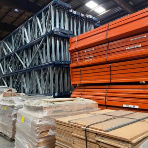 Stacks of industrial racking components and materials are stored in a warehouse. On the left, there are large blue and silver trusses neatly piled. In the center, bundles of wrapped wooden boards are stacked on pallets. On the right, bright orange beams are arranged in a tall pile, secured with black straps. The warehouse ceiling is visible, with natural light filtering through skylights, creating a well-lit storage area. The organized setup indicates efficient use of space and readiness for assembly or distribution.