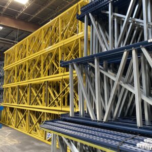 A spacious warehouse containing large stacks of metal pallet rack shelving frames in various colors, including yellow, blue, and gray. The pallet rack frames are neatly arranged and piled high, showcasing an organized inventory. The warehouse features a high ceiling with industrial lighting, highlighting the structural details of the shelving units