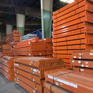 A large warehouse with stacks of orange metal beams and other shelving materials neatly organized. The beams are arranged in various sizes and are stacked up high, indicating a well-stocked inventory. The ceiling is high, with industrial lighting illuminating the space.