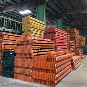 A warehouse interior features neatly stacked piles of metal beams and components organized by color. The stacks include orange, yellow, red, green, and blue beams, indicating a well-structured and efficient storage system. The beams are labeled for easy identification and retrieval. The warehouse has a high ceiling with exposed beams and industrial lighting, providing ample illumination. The concrete floor is clean and spacious, allowing for easy movement and access to the stored materials. The overall scene showcases a highly organized storage area, ready for construction or assembly operations.