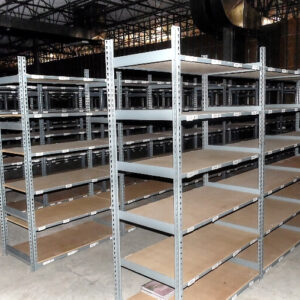 The image shows a large warehouse area filled with rivet shelving units. These shelving units are made of metal with particle board shelves. The shelves are arranged in rows and columns, providing ample storage space for various items. Each unit has multiple adjustable shelves supported by metal uprights with rivet holes, allowing for flexibility in shelf height. The rivet shelving units are designed to be sturdy and durable, suitable for heavy-duty storage needs. The warehouse environment features a high ceiling with industrial lighting, and the concrete floor is typical of such settings. In the background, there is an office chair, indicating a workspace within the storage area. The overall setup highlights the organization and efficient use of space in the warehouse.