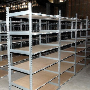 A large, indoor warehouse area is filled with rows of empty metal rivet shelving units. The shelves are constructed from grey metal frames with particleboard shelving surfaces. The units are arranged in a grid-like pattern, providing numerous aisles for storage and organization. The concrete floor is visible, and the warehouse has high ceilings with exposed beams and industrial lighting. In the background, a blue office chair on wheels is positioned near one of the shelving units, adding a touch of color to the otherwise industrial setting. The overall scene suggests a well-organized space, ready for stocking with inventory.