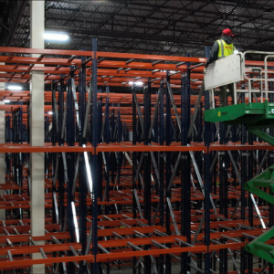 A worker stands on a green scissor lift, wearing a red hard hat and a yellow safety vest, high above the ground in a large warehouse. The scissor lift is elevated to allow the worker to reach the top levels of an extensive pallet rack shelving system. The pallet rack shelves have blue vertical frames and orange horizontal supports, creating a grid-like structure that stretches deep into the background. The warehouse ceiling is high, with industrial lighting illuminating the area. The scene highlights the process of installing or inspecting the high-rise shelving units, emphasizing the scale and height of the storage infrastructure.