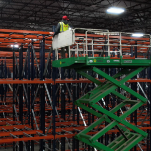 A worker stands on a green scissor lift, wearing a red hard hat and a yellow safety vest, high above the ground in a large warehouse. The scissor lift is elevated to allow the worker to reach the top levels of an extensive pallet rack shelving system. The pallet rack shelves have blue vertical frames and orange horizontal supports, creating a grid-like structure that stretches deep into the background. The warehouse ceiling is high, with industrial lighting illuminating the area. The scene highlights the process of installing or inspecting the high-rise shelving units, emphasizing the scale and height of the storage infrastructure.