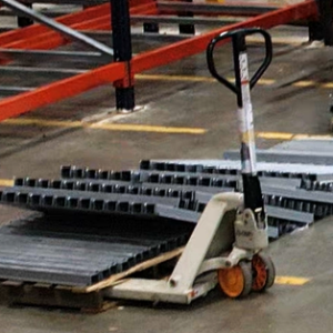 A warehouse scene depicts the process of setting up or rearranging shelving units. In the foreground, a manual pallet jack is loaded with metal beams, neatly stacked on a wooden pallet. Adjacent to the pallet jack, more metal beams are laid out on the floor in organized rows. The concrete floor is marked with yellow lines, indicating designated pathways and storage areas, contributing to an organized and safe work environment.