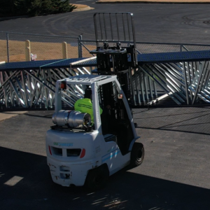 A forklift is shown outdoors, lifting a large, metallic truss structure. The forklift operator is wearing a bright green shirt. The forklift is labeled "UniCarriers" on its side and appears to be powered by a propane tank mounted at the back. The truss spans the width of the image, and the scene is set on a paved surface with a chain-link fence and a parking area in the background.