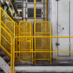 A yellow industrial handrail system is installed on a metal platform. The handrail features multiple vertical and horizontal bars with mesh panels for added safety. The setup includes a staircase leading up to the platform, with the handrails providing protection on both sides and around the elevated area. The overall environment appears to be within a factory or industrial facility, indicated by the machinery and equipment in the background.