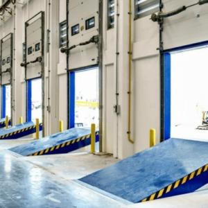 A modern warehouse with a row of loading dock doors, each equipped with blue dock levelers featuring yellow and black safety markings. The dock levelers are positioned to facilitate easy loading and unloading of goods from trucks. The interior space is clean and well-lit, with polished concrete floors and yellow safety bollards near each dock door. The overall setup highlights an organized and efficient material handling area, ready to accommodate large volumes of shipments.