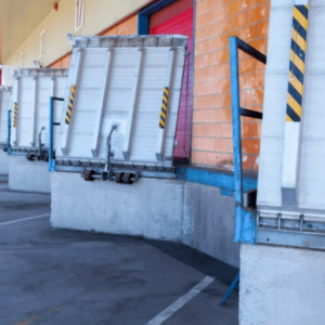 A row of loading dock doors with raised dock levelers at an industrial facility. Each dock leveler is positioned to bridge the gap between the dock and a truck bed, featuring yellow and black safety markings. The setting includes a concrete platform with blue metal railings and red dock doors, suggesting an active area for loading and unloading goods. The scene highlights the infrastructure in place to support efficient and safe material handling operations.