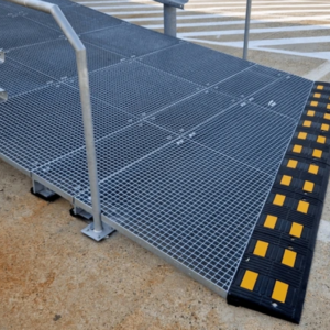 A metal dock ramp with a grated surface provides a stable platform for loading and unloading. The ramp features a yellow and black edge for increased visibility and safety. It is equipped with handrails on the sides for additional support and safety. The ramp is set up on a concrete surface, indicating it is part of a loading dock area. The design ensures durability and efficient use in industrial settings.