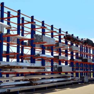 A cantilever pallet storage rack with blue vertical supports and orange horizontal arms holding various metal beams and profiles, located outdoors under a clear blue sky. Some materials are stacked neatly on the racks, while others are wrapped in protective covers. The scene is set in an industrial area with a white building in the background and trees visible in the distance.