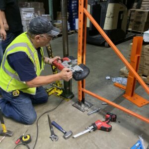 A worker wearing a yellow safety vest and a cap is kneeling on the floor of a warehouse, using a power tool to repair orange metal pallet rack upright. Various tools, including hammers, wrenches, and a measuring tape, are scattered around him. Behind the worker, there are pallets of boxes and other warehouse equipment. The scene depicts a pallet rack repair process, highlighting the hands-on work involved in maintaining warehouse infrastructure.