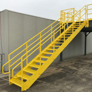 Exterior view of a bright yellow metal staircase with handrails leading up to an elevated platform. The staircase is attached to the side of a building with a gray exterior wall. The steps are perforated for traction, and the structure appears sturdy and well-constructed. The surrounding area is paved and clean, indicating a well-maintained facility.